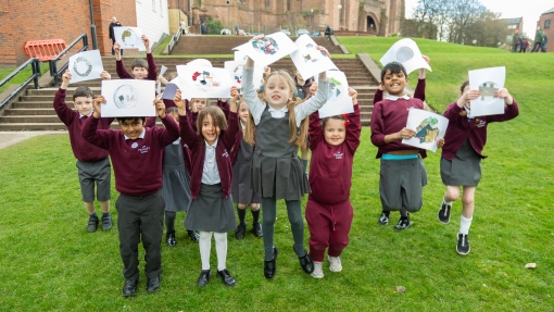Students on the grass holding up art work, standing in front of a red brick building 