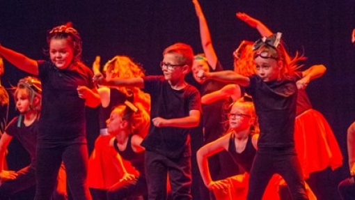 A group of children dance on a stage in dark red lighting. 