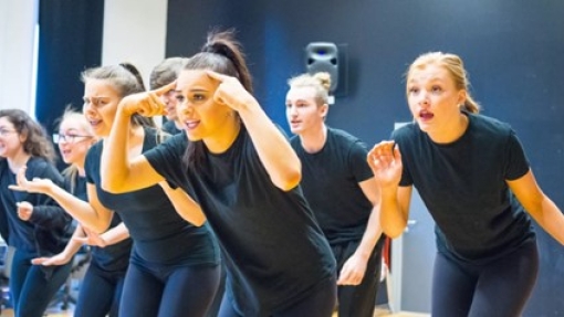 A group of young women stand on a stage, staring at an audience with wide eyed and expressive looks on their faces. They are wearing black tops with a blue bricked wall behind them. 