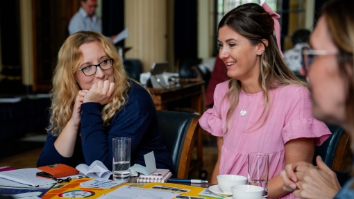 Two white females sitting at a table with documents and post-it notes having a discussion. One lady is looking thoughtful with her hand on her chin. 