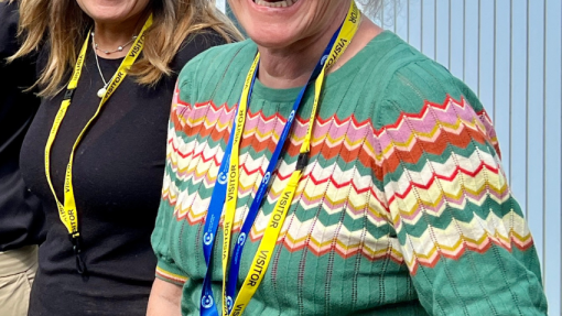 A woman with grey hair and a bright green top is looking to the camera, smiling. Another woman is smiling behind her.