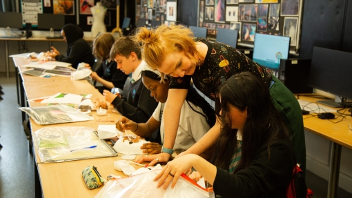 Pupils sat at a long desk and a teacher is leaning over one of the pupils, pointing at their work.
