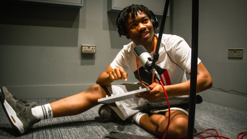 A young person sitting on the floor with a microphone in front of him and he's holding a piece of technical equipment.