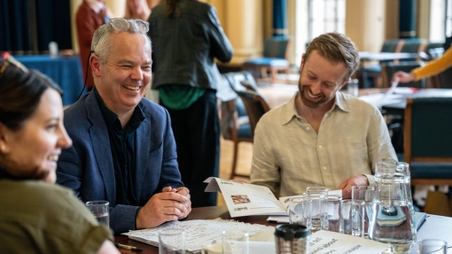 Two men sitting at a table in a meeting room, one is looking down at a document, the other is looking up, smiling.