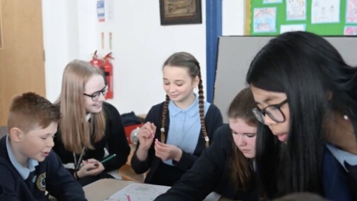 A group of school children seated around a table in a classroom, working on worksheets with colored pencils. They are wearing school uniforms, and their faces are blurred for privacy. In the background, a green bulletin board displays various artworks, and a fire extinguisher is mounted on the wall.