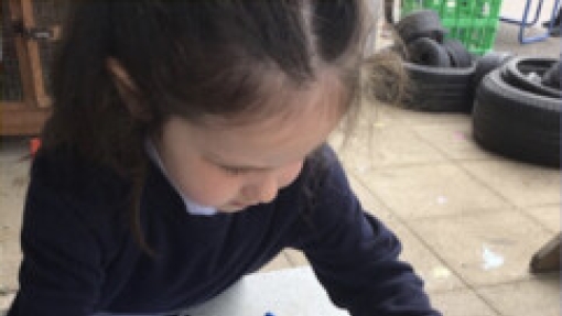 A child face measures an object on a tray using a yellow and green striped ruler. The child wears a dark long-sleeve shirt. On the table are blue gloves, a magnifying glass, and papers. Tires and other outdoor items are visible in the background.