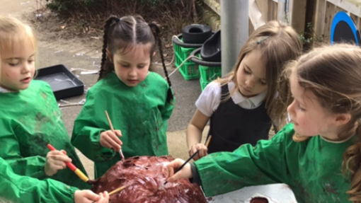 A group of children wearing green aprons are gathered around a table, painting a large, brown, dome-shaped object. They use brushes and small containers of paint. 