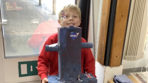 A child in a red sweater holds a homemade space rover model painted dark blue with a NASA logo. They stand in front of a door with glass panels. A desk with papers and items is visible to the right.