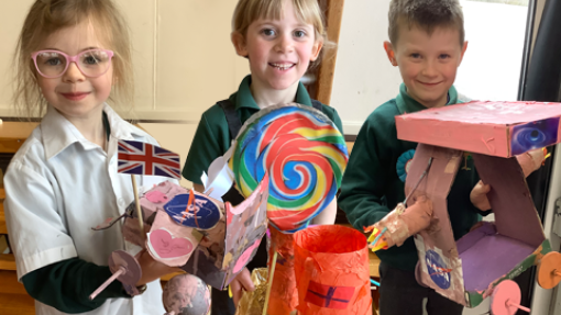 Three children stand side by side, each holding a colorful craft project. The child on the left holds a model featuring a Union Jack flag and NASA logo. The middle child displays a large, multicolored lollipop decoration. The child on the right holds a pink and purple model resembling a vehicle or structure. 