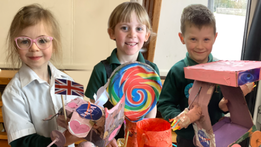 Three children standing side by side, each holding a colorful handmade craft. The child on the left holds a project featuring a small Union Jack flag and a NASA logo. The middle child holds a large, multicolored lollipop-shaped object. The child on the right holds a pink and purple craft resembling a vehicle with wheels.