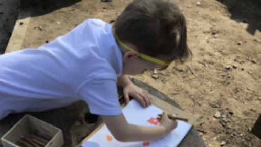 A child in a white shirt and yellow headband lies on the ground, drawing on paper attached to a clipboard. Colored pencils are visible in an open box nearby. The ground is dirt with small rocks and wooden planks scattered around.