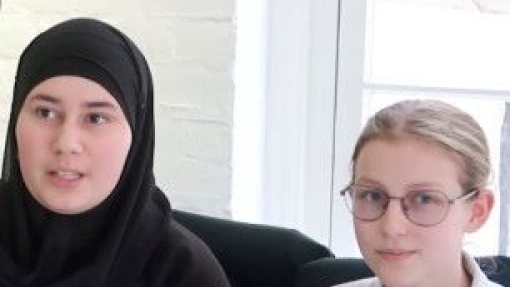 Two individuals seated side by side on a couch. The person on the left wears a black hijab and a black top with the word 'DANCE' and an emblem. The person on the right wears glasses and a white polo shirt with black trim, featuring an emblem and the text 'PE'