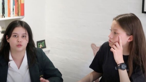 Two individuals seated in a room with white walls. The person on the left wears a dark blazer over a white shirt; the person on the right wears a black t-shirt and a watch on their left wrist.