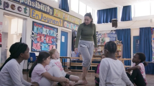 Children sit on a classroom floor around a standing adult. Faces are pixelated for privacy. The room features colorful posters with words like 'Responsibility' and 'Enjoyment,' blue curtains, and a bookshelf in the background.