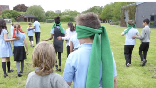 A group of children participating in an outdoor activity on a grassy field. Some children wear green fabric blindfolds while others guide them by holding their shoulders or arms. They are dressed in school uniforms with light blue shirts and dark bottoms. In the background, there is an ivy-covered building and several trees.