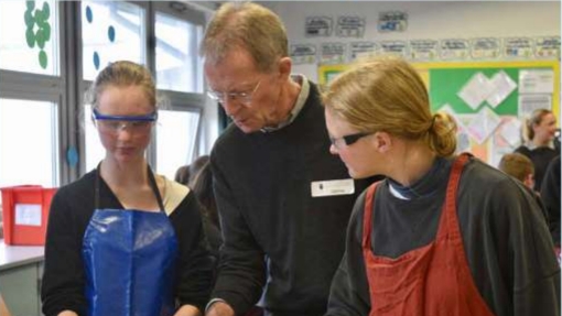 In a classroom, a teacher wearing a black sweater demonstrates something on a device placed on a table. Two students observe closely—one in a blue apron holding a ruler, and the other in a red apron looking at the device.