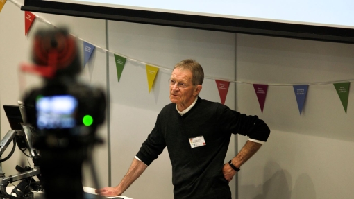 A person stands at a podium in a lecture hall, one hand on the podium and the other on their hip. Behind them, colorful triangular flags are strung across the wall. An out-of-focus camera in the foreground appears to be recording or streaming the event.