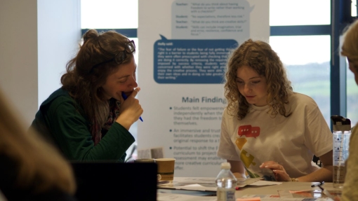 Two people sit at a table engaged in discussion or study. One wears a green sweater and holds a pen; the other wears a white t-shirt with speech bubble graphics. The table is scattered with papers, notebooks, a water bottle, and coffee cups.