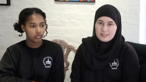 Two individuals seated at a table, both wearing black long-sleeve shirts with a white logo on the left chest that reads 'DANCE' and includes an image of a building. The person on the left has curly hair tied back and rests one hand on the table; the person on the right wears a headscarf. Behind them is a white brick wall with framed artwork, including a colorful abstract painting in the centre.