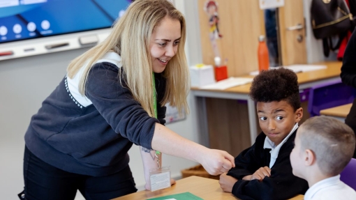 A teacher with long blonde hair, wearing a black and white sweater, stands beside a desk in a classroom, handing an item to a seated student with short curly hair in a black sweater and white shirt. Another student in a white shirt sits nearby. The desk holds papers and a green notebook, with additional desks, chairs, and classroom supplies visible in the background.