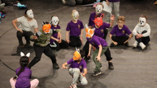 Children perform on stage wearing animal masks, including foxes and pandas. They are dressed in casual clothing, with some standing and others sitting or crouching in a circle. In the background, more children are seated, watching the performance.