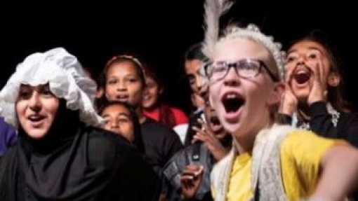 A group of people in costume, including yellow shirts, black outfits, a white bonnet with a black shawl, and a feathered headpiece with a fur vest. Faces are blurred for privacy. The dark background suggests an indoor or stage setting.