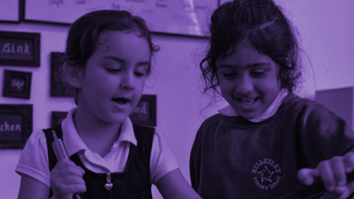 Two children stand in a classroom. One holds a tool and wears a white shirt with a dark pinafore; the other wears a dark sweatshirt with a 'Stella Maris Primary School' emblem.