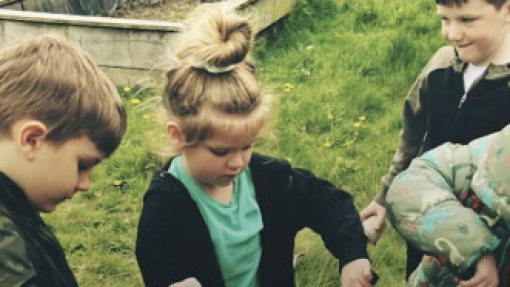 A group of children gathered outdoors on a grassy area, participating in a gardening activity. One child in the center is using a small shovel to dig into the soil.
