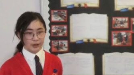 A student in a red cardigan and tie stands in front of a classroom display board. The board features open book images with text and small photos, bordered by a scalloped edge. On the table in front are various models or projects, including a pyramid and rectangular structures.