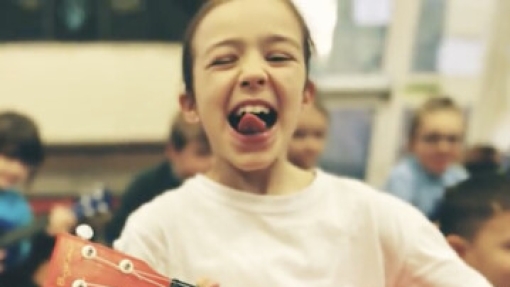 A person in a white shirt holds a red four-string guitar-like instrument with tuning pegs. Other people are visible in the background of a room with large windows.