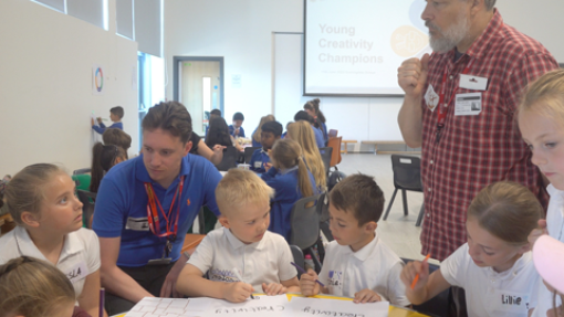 A classroom with children seated at tables, working on large sheets of paper using markers. Two adults—one in a blue shirt and one in a red checkered shirt—are assisting them. A projector screen at the front displays the text 'Young Creativity Champions.' Natural light enters through tall windows on the left.