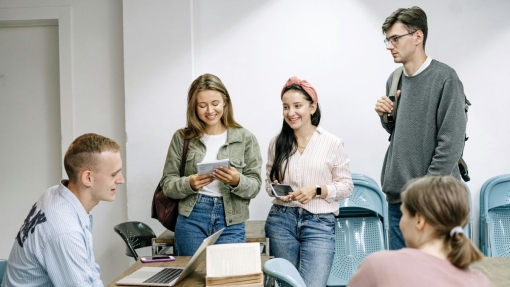 Five people in a classroom. One seated at a desk with a laptop and books, while four others stand nearby, two holding notebooks or papers. Blue chairs are arranged in rows, with a white wall in the background.