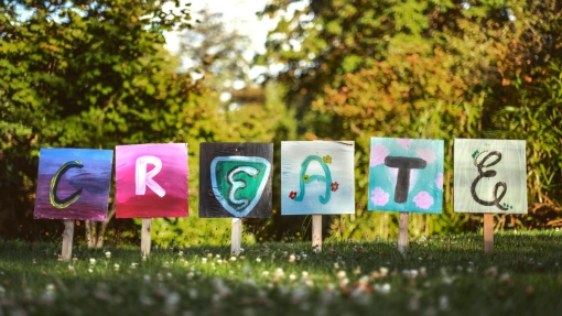 Six colorful signs on a grassy field spell out 'CREATE,' each letter painted uniquely on square canvases mounted on wooden stakes, with lush green trees in the background.