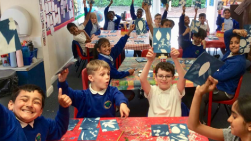 Primary school children in blue uniforms sit at red-covered tables in a classroom, proudly holding up blue and white paper cut-out art projects. Art supplies are scattered on the tables, and the background features colorful wall decorations and sunlit windows.