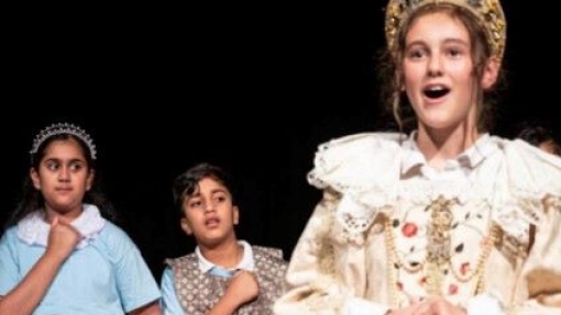 Three individuals stand on a stage. The person on the right wears an elaborate historical costume with lace, while the other two wear simpler outfits. A dark background highlights the figures.