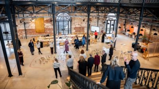 Interior of a spacious, industrial-style venue with high ceilings, exposed brick walls, and large windows. People are engaged in activities and conversations around tables and displays. The scene is viewed from a staircase with metal railings in the foreground.