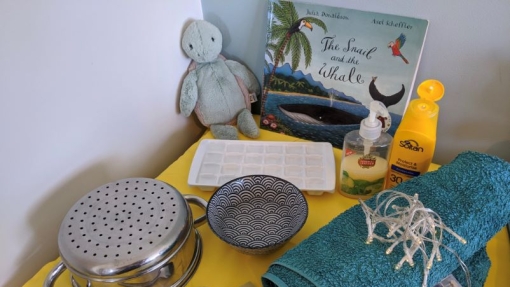A yellow table displays a collection of items: a green stuffed toy, a children's book titled 'The Snail and the Whale,' an ice cube tray, a bottle of hand soap, sunscreen lotion, a teal towel with string lights draped over it, a small metal colander with handles, a patterned bowl, a spoon and fork set, and two small rectangular objects