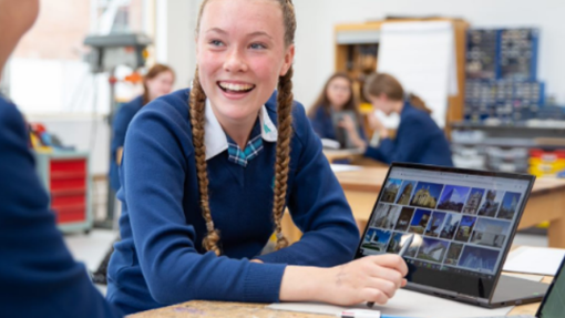 A student with braided hair in a blue school uniform sits at a classroom desk, viewing images of buildings and architecture on a laptop. Other students in similar uniforms are visible in the background, engaged in various activities.
