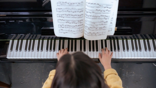 Overhead view of a person in a yellow sweater playing the piano, with their hands on the keys and an open sheet music book on the music stand displaying multiple lines of musical notation.