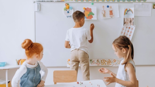Three children in a classroom: one stands at a whiteboard drawing or writing, while two others sit at a table working with small objects and papers. The whiteboard displays colorful drawings and numbers from 5 to 10.