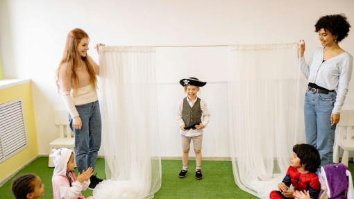 "Indoors on green carpeted flooring, two adults hold up sheer white curtains to form a makeshift stage. A child dressed as a pirate stands center stage, while four other children in costumes—including animal onesies and superhero outfits—sit on the floor watching and clapping.