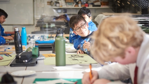 A student wearing glasses and a blue overall is painting and smiling in a classroom