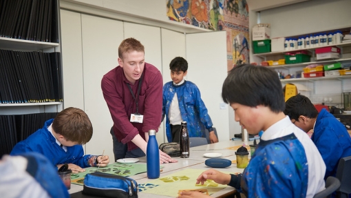 A teacher in a burgundy shirt leans in to help a group of students sitting at a table with their painting.
