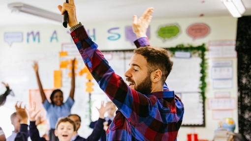 A teacher at the front of a classroom with his arms raised in the air. There are children in the background with their hands also in the air. 