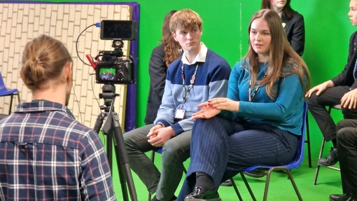 People seated in front of a green screen being filmed by a camera on a tripod.