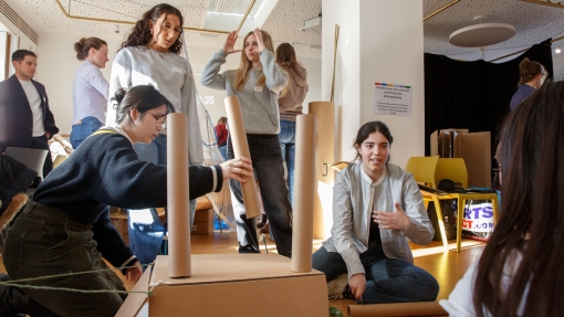 Group of people engaged in a hands-on creative activity indoors, building structures with cardboard tubes and blue blocks, with others standing and observing in the background.
