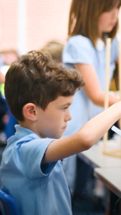 In a classroom, two children in light blue shirts work together on a project using wooden frames and plastic sheets. One holds a hot glue gun while the other positions the frame. Other students are engaged in similar activities in the background.