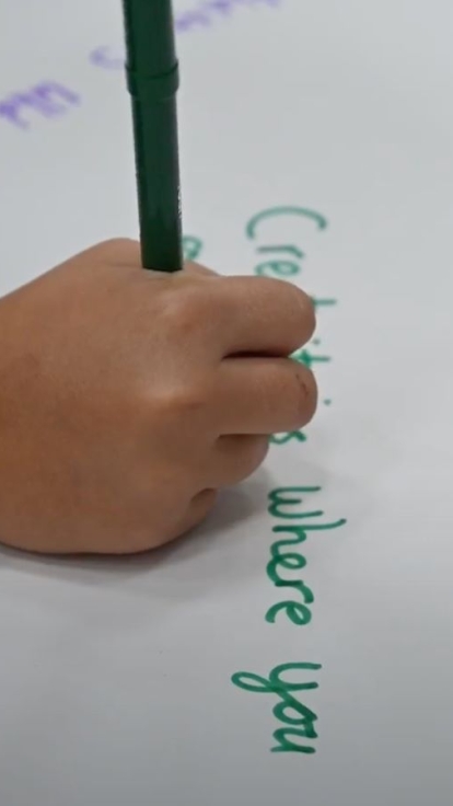 Close-up of a child writing with a green marker on white paper, wearing beaded bracelets. Colorful handwritten words like 'Creating' and 'exploring new things' are visible.