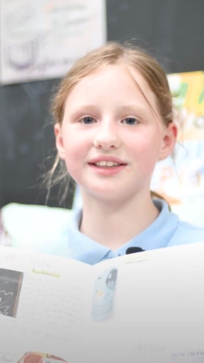 A child holds an open book displaying handwritten notes and ocean-themed illustrations. In the background, a blackboard and a shelf with colorful books, including 'Here Comes Sophie' by Dick King-Smith, are visible.