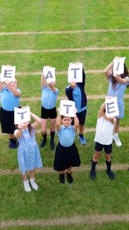 A group of children standing on a grassy field, arranged in three rows. Each child holds a white card with a black letter, collectively spelling out 'CREATIVITY MATTERS.' The children are dressed in blue and white clothing.